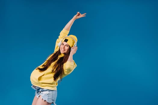 A pregnant girl in a yellow jacket and headphones stands on a blue background.