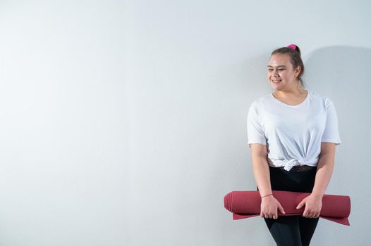 Young fat caucasian woman holding a sport mat. Charming plus size model in sportswear stands on a white background.