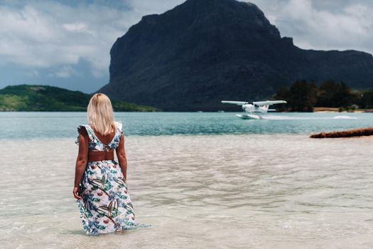 A girl in a swimsuit stands in the ocean and waits for a seaplane against the background of mount Le Morne on the island of Mauritius.A woman in the water looks back at a plane landing on the island of Mauritius.