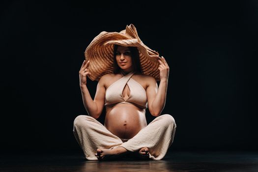 a pregnant woman in a straw hat sits on the floor in beige clothes in a studio on a black background.