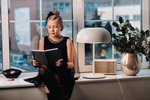 A stylish girl in a black dress sits on the windowsill by the window with a book in her hands.