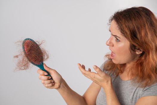 Caucasian woman with a grimace of horror holds a massage comb with a bun of hair. Hair loss and female pattern baldness