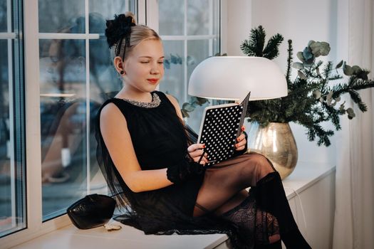 A stylish girl in a black dress sits on the windowsill by the window with a book in her hands.