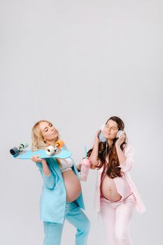 Two pregnant girls in turquoise and pink suits with glasses of juice, a skate and headphones stand on a gray background.