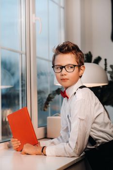 At the window stands a boy in a shirt with a red bow tie and glasses, holding a red book in his hands.