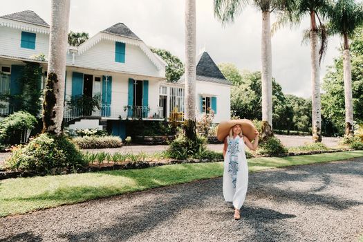 Young beautiful sexy woman on a tropical island. Stylish girl in a white long dress and a large straw hat. Summer holidays in Mauritius happiness .Against the background of a colonial house.