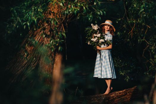 A stylish woman with a straw hat and bare feet poses on a fallen tree with a bouquet of lilacs in a Sunny spring Park. A quiet portrait of a beautiful girl standing in a purple lilac .