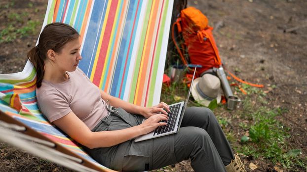 Caucasian woman working on laptop while sitting in a hammock in the forest. Girl uses a wireless computer on a hike