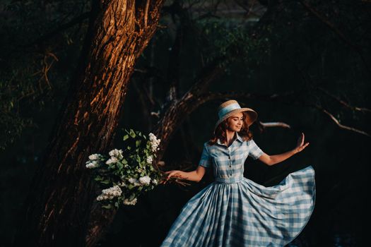 Portrait of a beautiful girl with long hair, a straw hat and a long summer dress with lilac flowers in the garden.