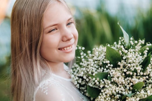 A beautiful nine-year-old blonde girl with long hair in a long white dress, holding a bouquet of lilies of the valley flowers, walking in nature in the Park.Summer, sunset