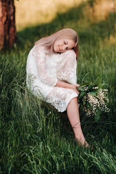 A beautiful nine-year-old blonde with long hair in a long white dress, holding a bouquet of lilies of the valley, sits on a stone in the Park.Summer, sunset