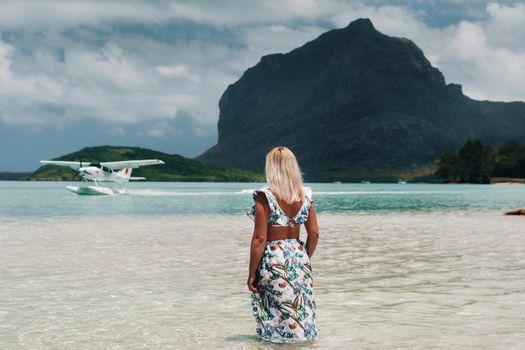 A girl in a swimsuit stands in the ocean and waits for a seaplane against the background of mount Le Morne on the island of Mauritius.A woman in the water looks back at a plane landing on the island of Mauritius.