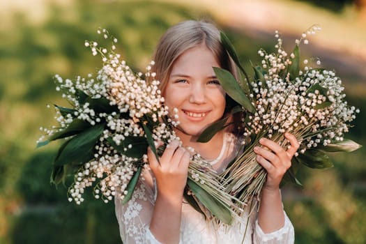 A beautiful nine-year-old blonde girl with long hair in a long white dress, holding a bouquet of lilies of the valley flowers, walking in nature in the Park.Summer, sunset