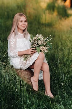 A beautiful nine-year-old blonde girl with long hair in a long white dress, holding a bouquet of lilies of the valley flowers, walking in nature in the Park.Summer, sunset