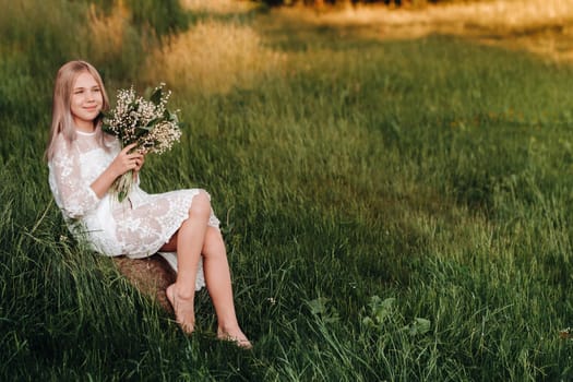 A beautiful nine-year-old blonde girl with long hair in a long white dress, holding a bouquet of lilies of the valley flowers, walking in nature in the Park.Summer, sunset