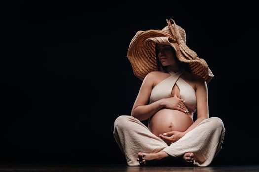 a pregnant woman in a straw hat sits on the floor in beige clothes in a studio on a black background.