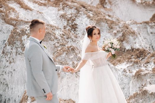 A beautiful couple of lovers posing in a white salt mountain. A young woman in a stylish wedding dress and a beautiful stylish man in a gray suit. The concept of the wedding day