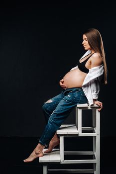 pregnant woman sitting on a ladder chair in a studio on a black background.