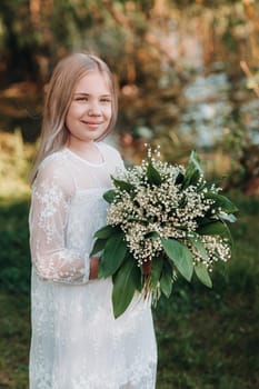 A beautiful nine-year-old blonde girl with long hair in a long white dress, holding a bouquet of lilies of the valley flowers, walking in nature in the Park.Summer, sunset