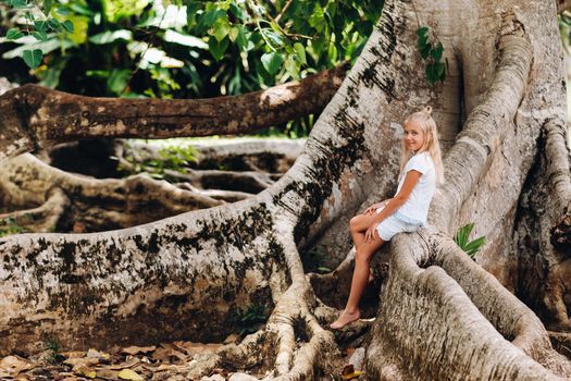 Summer portrait of a happy little girl on the island of Mauritius sitting on a huge tree.A girl sits on a large tree in the Botanical garden of the island of Mauritius.
