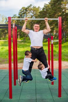 Caucasian man and two boys doing exercises outdoors. The father pulls himself up on the horizontal bar with his sons on the playground