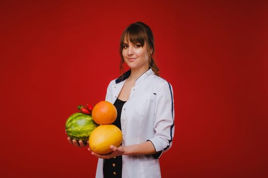 a female doctor nurse in a white coat with fruit in her hands poses on a red background, melon, watermelon, strawberry and grapefruit.