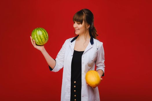 a female doctor nurse in a white coat with fruit in her hands poses on a red background, melon, watermelon