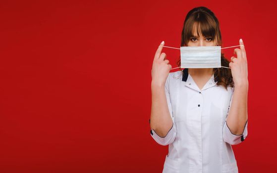 A female doctor stands on a red background and holds a medical mask in her hands.