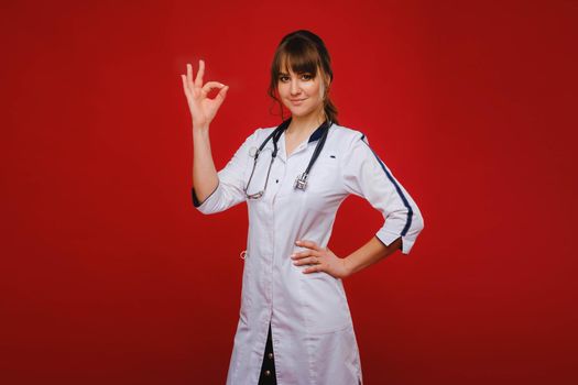 A young doctor stands on a red background and shows his hand everything will be fine. The doctor shows OK