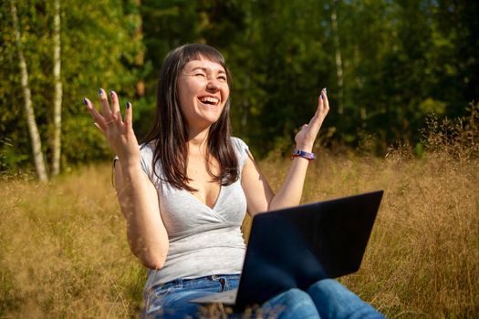 happy young woman with laptop on a sunny lawn. freelancer works in nature. student studies remotely on nature landscape outdoor