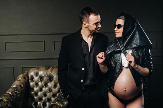 a pregnant woman in black clothes and a headscarf and a man in a suit in a studio on a dark background.