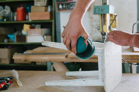 A man is polishing a white old stool. Grinding machine. Restoration of furniture.