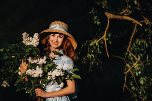 Portrait of a beautiful girl with long hair, a straw hat and a long summer dress with lilac flowers in the garden.