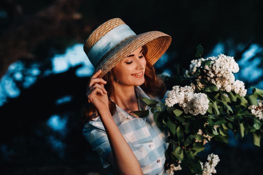 Portrait of a beautiful girl with long hair, a straw hat and a long summer dress with lilac flowers in the garden.