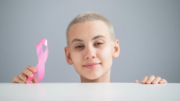 Young woman with short hair wearing a white t-shirt holding a pink ribbon as a symbol of breast cancer on a white background