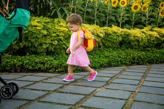 adorable infant girl in pink dress with baby backpack walk the road in park side view