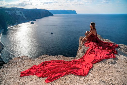 Side view a Young beautiful sensual woman in a red long dress posing on a rock high above the sea during sunrise. Girl on the nature on blue sky background. Fashion photo.