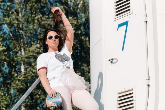 A girl in white clothes and glasses with a skate in her hands is photographed near large wind turbines in a field with trees.Modern woman with a riding Board in a field with windmills.