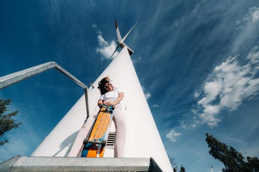 A girl in white clothes and glasses with a skate in her hands is photographed near large wind turbines in a field with trees.Modern woman with a riding Board in a field with windmills.