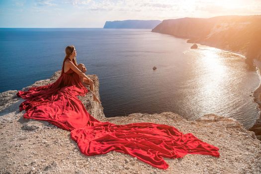 Side view a Young beautiful sensual woman in a red long dress posing on a rock high above the sea during sunrise. Girl on the nature on blue sky background. Fashion photo.