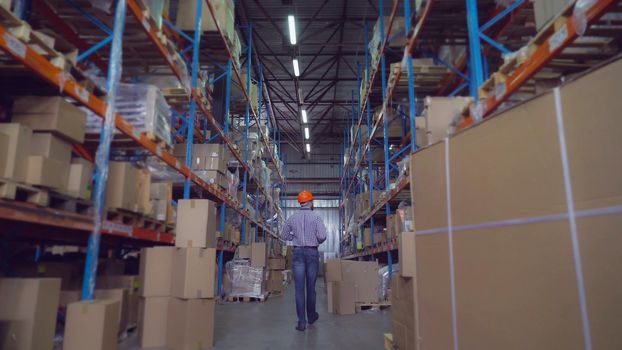 Manager at warehouse going along metal racks looking for some box check delivery. Rear back view. Man wearing casual shirt hard hat and jeans holding folder with document.