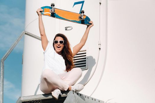 A girl in white clothes and glasses with a skate in her hands is photographed near large wind turbines in a field with trees.Modern woman with a riding Board in a field with windmills.