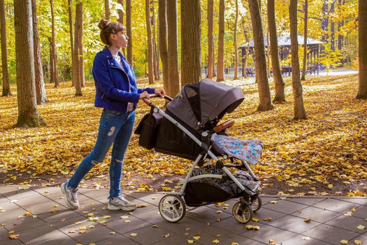 young mother goes with a stroller in the autumn park. a one-year-old infant is sleeping in a carriage. tired mom