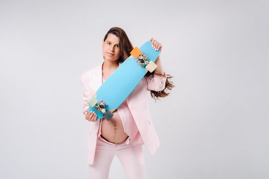 A pregnant girl in a pink suit with a skateboard in her hands stands on a gray background.