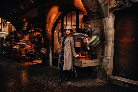 a girl in a coat and hat stands near baskets in the old town of Annecy, spending time outdoors exploring the European city. An elegant young lady in a coat and hat enjoys a view of the old town in France.