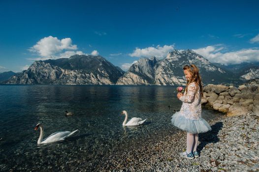 A little girl in a smart white dress walks along the embankment of lake Garda.A girl is photographed against the background of a mountain and lake in Italy.Torbole.