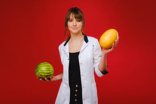 a female doctor nurse in a white coat with fruit in her hands poses on a red background, melon, watermelon