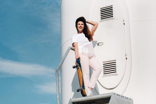 A girl in white clothes with a skate in her hands is photographed near large wind turbines in a field with trees.Modern woman with a Board for riding in a field with windmills.