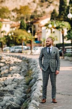 A bearded man in a strict grey three-piece suit stands against the backdrop of the Alps near Torbole,Italy.