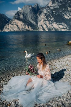 a girl in a smart white dress is sitting on the embankment of lake Garda.A woman is photographed against the background of a mountain and lake in Italy.Torbole.